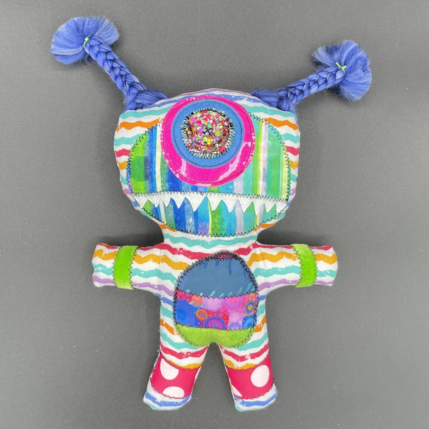 A colorful handmade fabric doll or plush toy with braided blue pigtails, a round embroidered face, and striped multicolor body panels, photographed against a plain gray background.