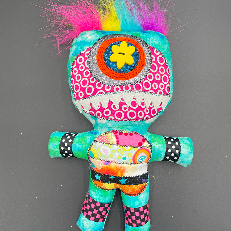 A colorful art doll with a monster character design, featuring a vibrant hair-like top with multi-colored patterns, and a stuffed body with decorative buttons and a flower detail on the head.
