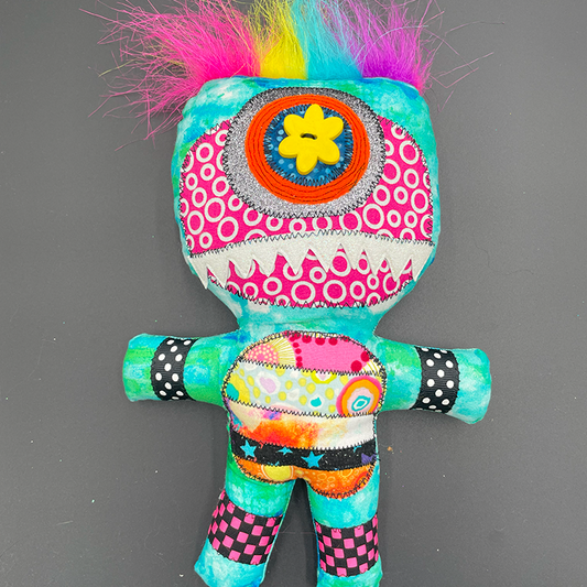 A colorful art doll with a monster character design, featuring a vibrant hair-like top with multi-colored patterns, and a stuffed body with decorative buttons and a flower detail on the head.