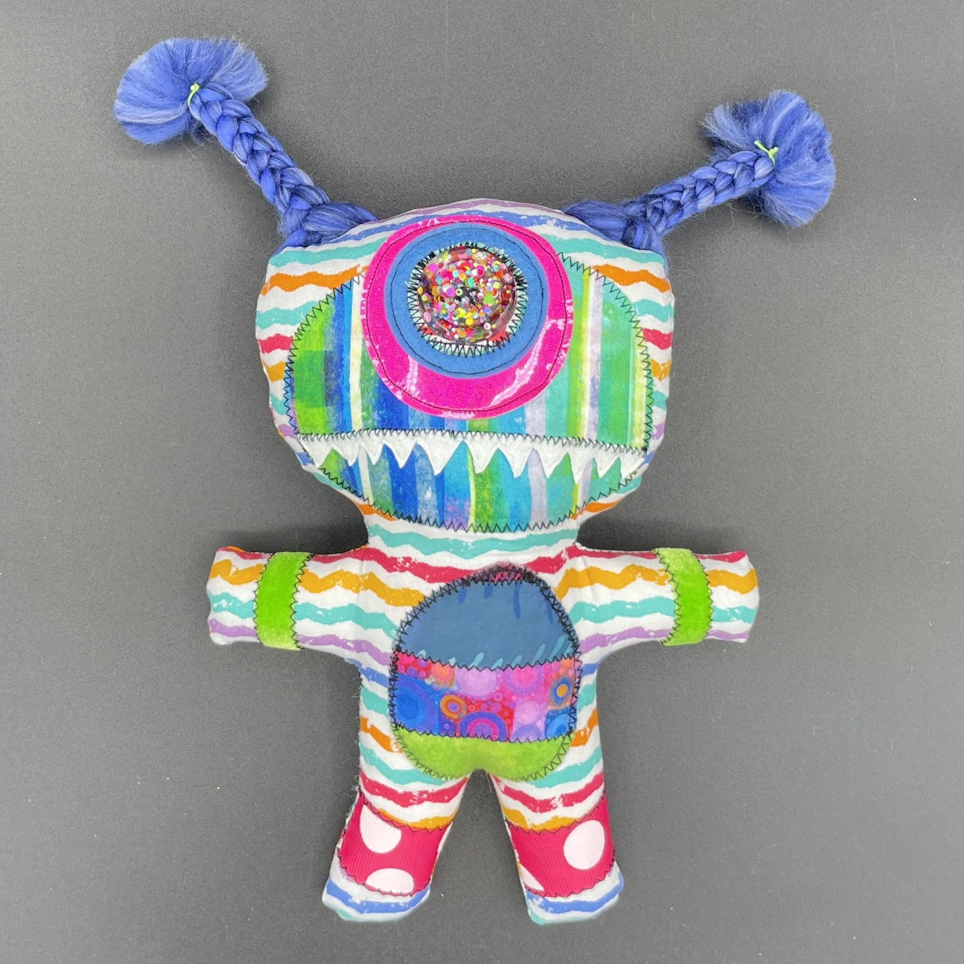 A colorful handmade fabric doll or plush toy with braided blue pigtails, a round embroidered face, and striped multicolor body panels, photographed against a plain gray background.